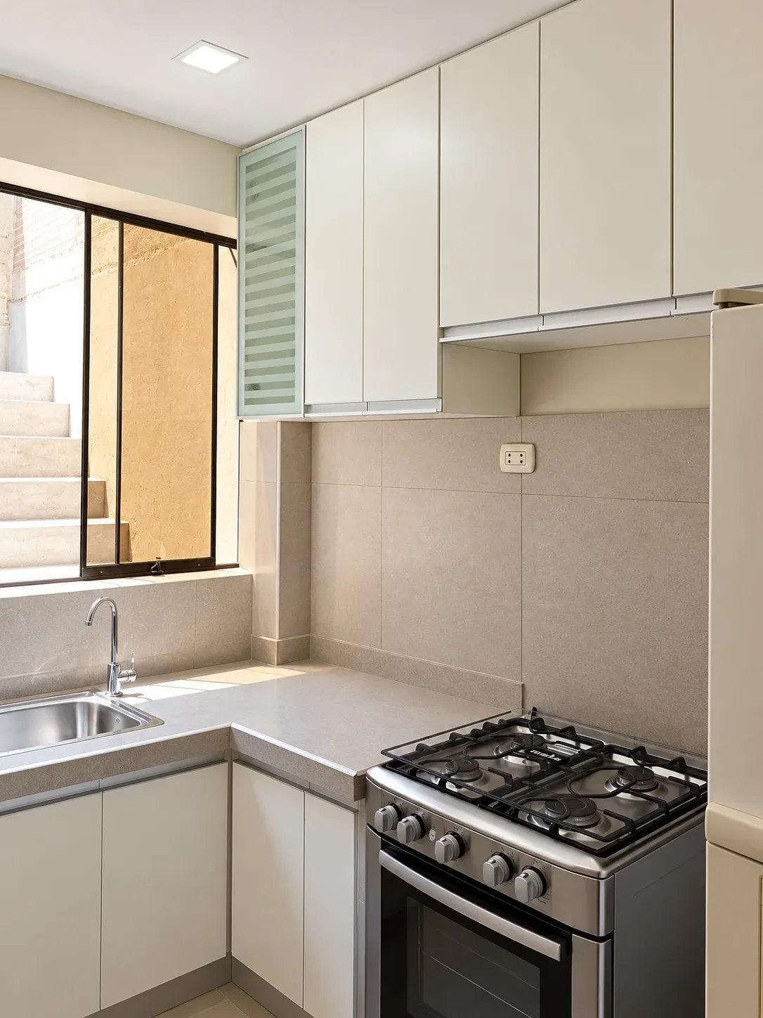 Cocina blanca en esquina con vitrina de franjas, lavadero bajo la ventana y cocina independiente.